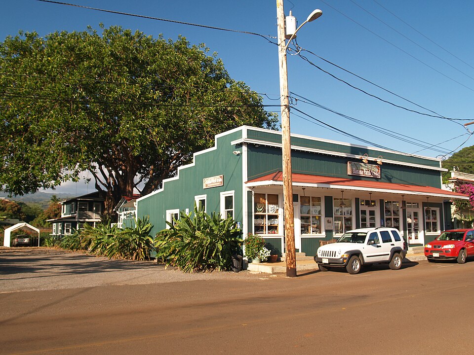 kauai art night Green building with a red roof, large tree, two parked cars, clear sky, and power lines.