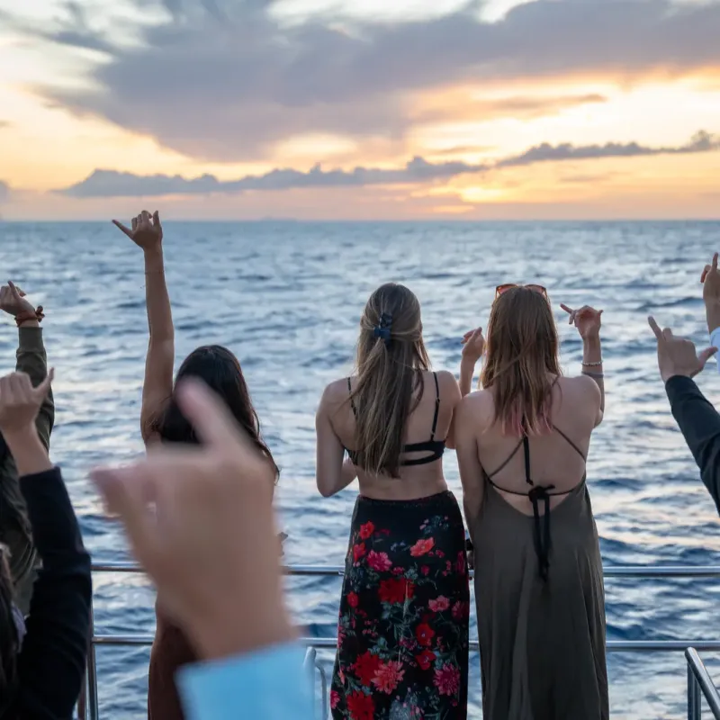 People on a boat raise hands in shaka gesture at sunset over the ocean.