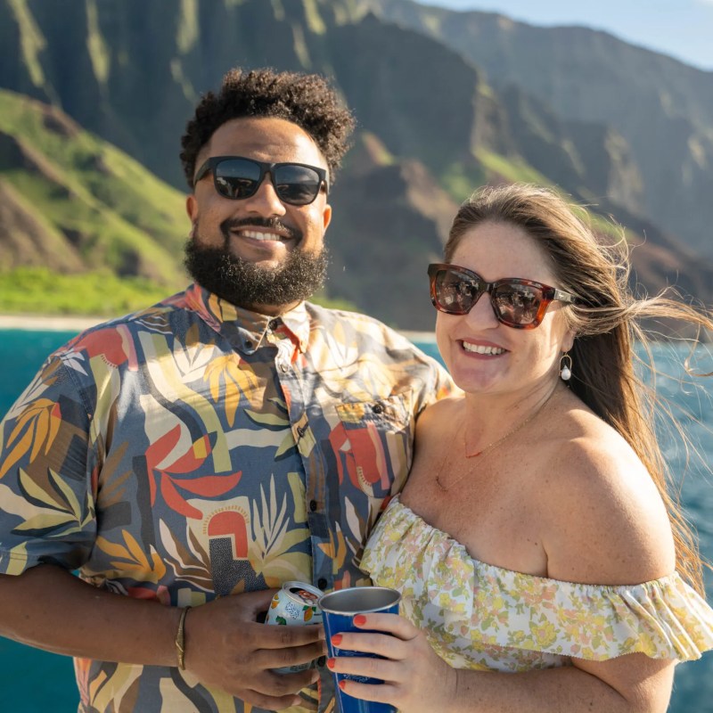 Smiling couple on boat, holding drinks, with mountains and ocean in background.