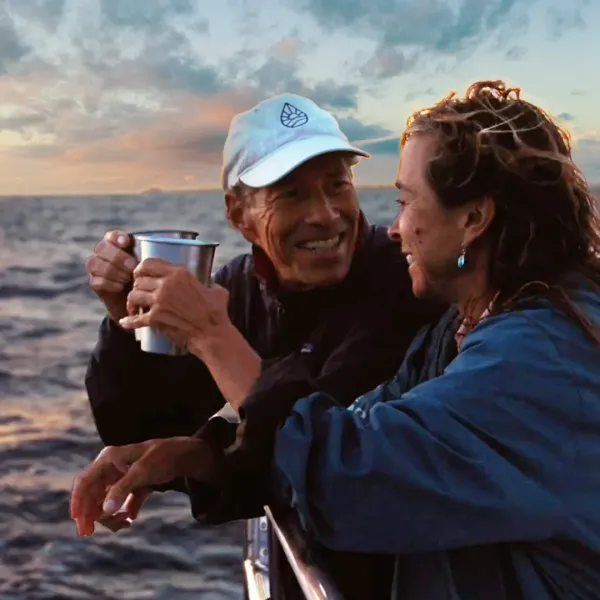 Two people smiling on a boat at sunset, one holding a metal cup.