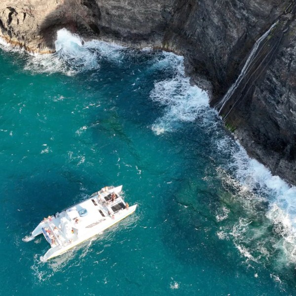 Aerial view of a catamaran near rocky cliffs with cascading waterfall.