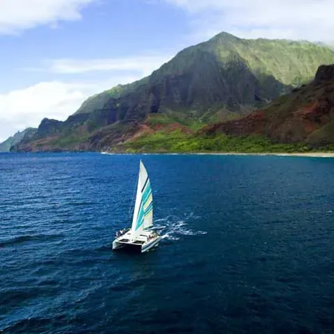 Sailboat with colorful sail on ocean near lush green mountain coastline.