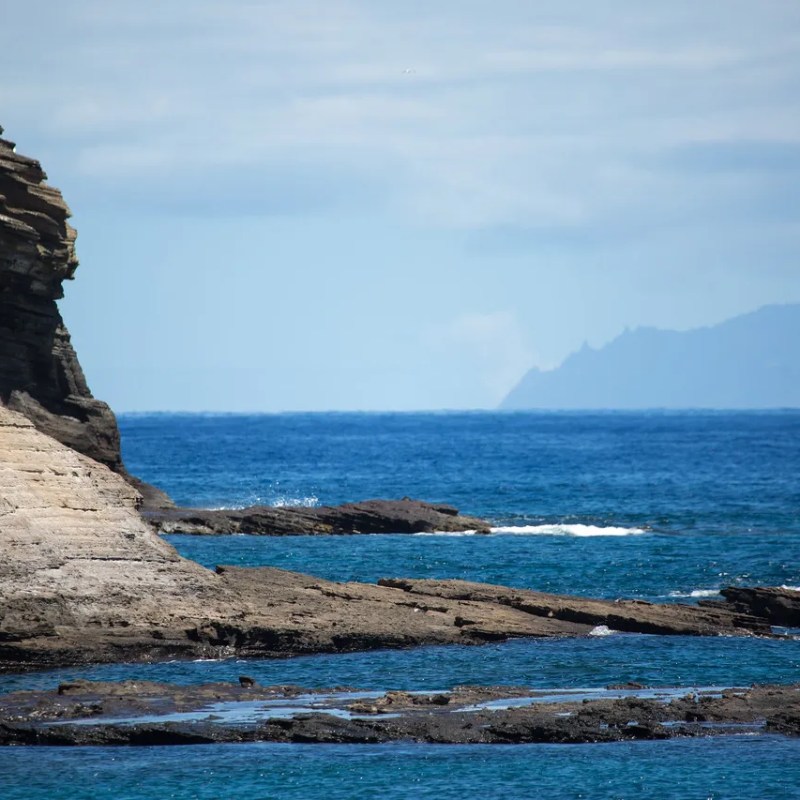Rocky cliffside by the ocean with distant mountains under a partly cloudy sky.
