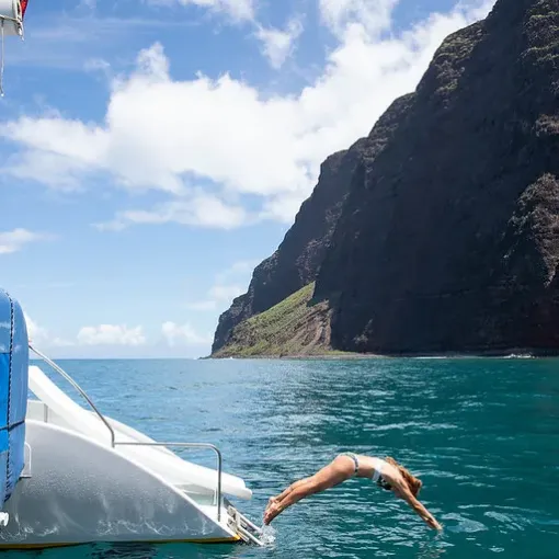 Person diving off a boat into clear blue ocean near a rocky cliff.