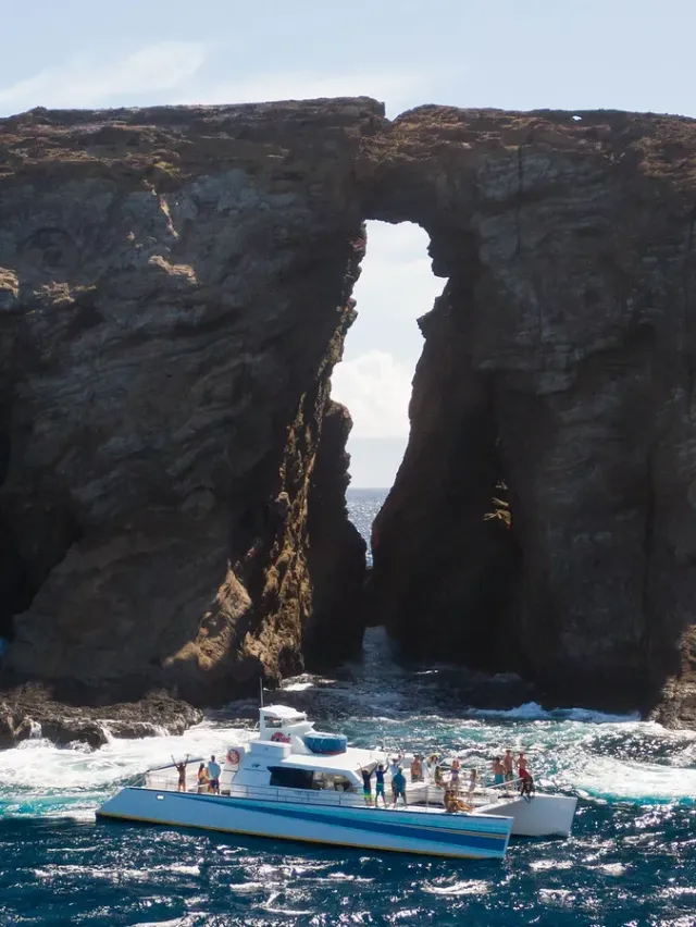 Boat near large rock arch with ocean waves and clear sky.