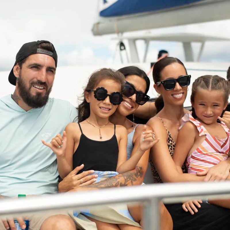 Group of adults and children posing together on a boat, smiling and wearing casual clothes and sunglasses.