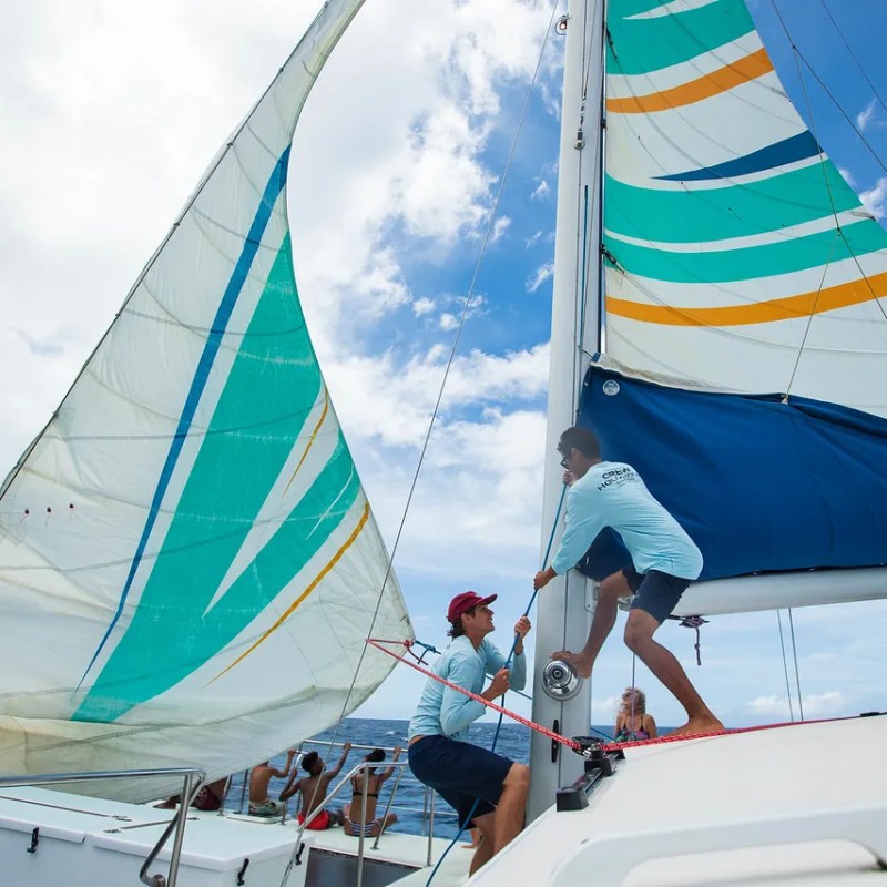 Two people adjusting sails on a boat under a partly cloudy sky.