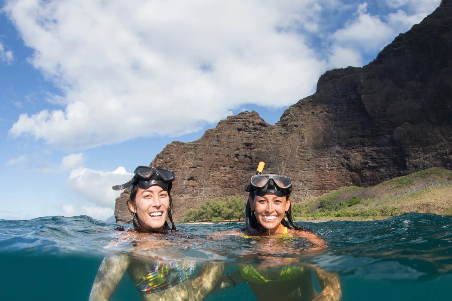 Two people snorkeling near rocky cliffs under a blue sky with clouds.