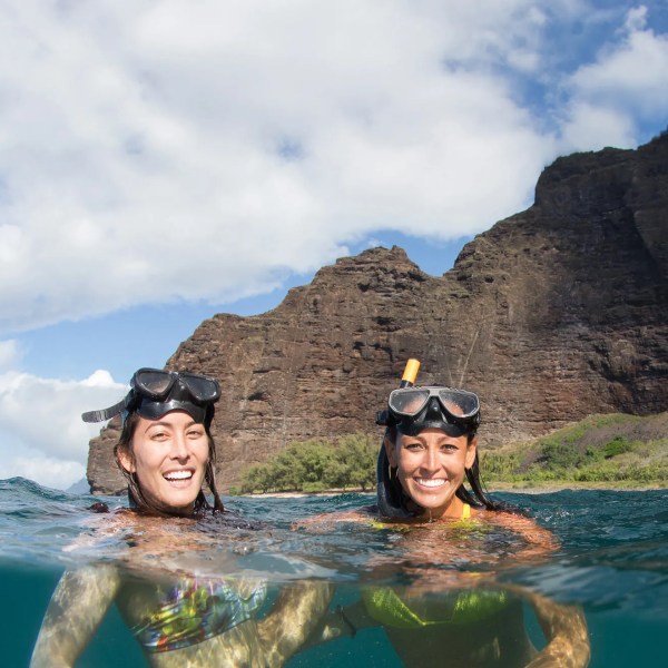 Two people snorkeling near rocky cliffs under a blue sky with clouds.