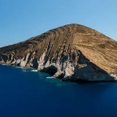 Aerial view of an island with rocky cliffs and dry grass, surrounded by deep blue ocean.