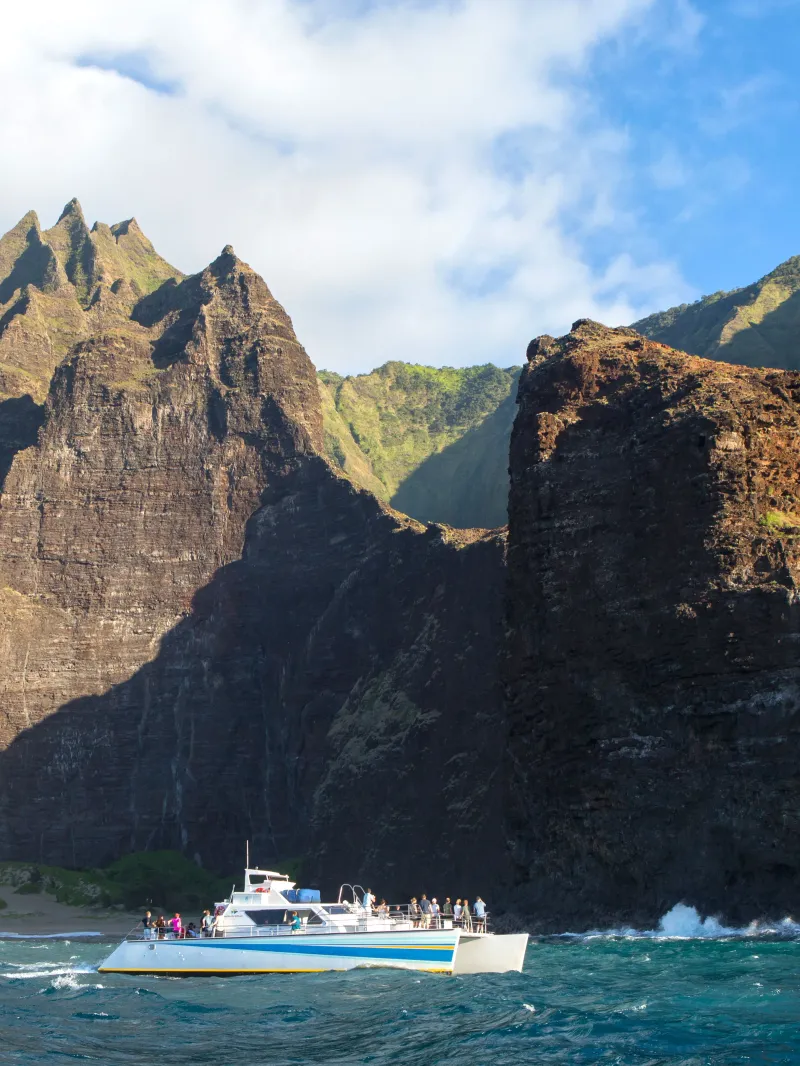 Boat sailing near towering cliffs with rugged terrain and green vegetation under a blue sky with clouds.