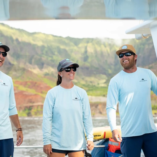 Three people in matching shirts and sunglasses smiling on a boat with a scenic mountain view.