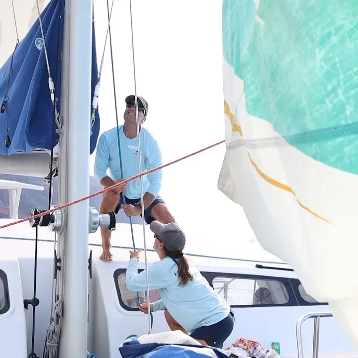Two people on a sailboat adjusting ropes with a mountainous coastline in the background.