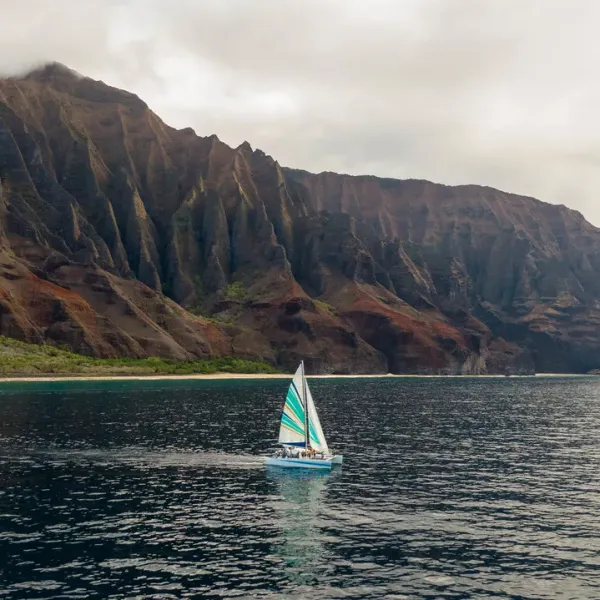 Sailboat on ocean near dramatic cliffs under cloudy sky.