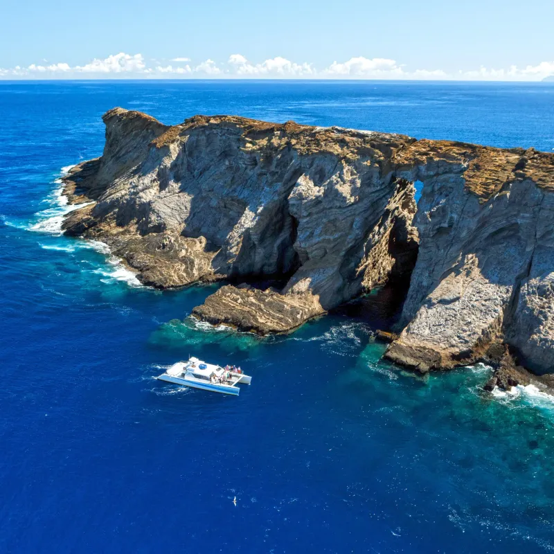 Aerial view of a boat near rugged cliffs and blue ocean under a clear sky.