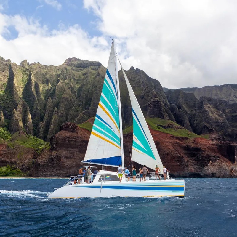 Catamaran sailing near steep, rugged cliffs with vibrant greenery and colorful sails.