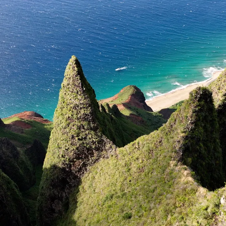 Aerial view of lush green jagged cliffs meeting a turquoise ocean and sandy beach.