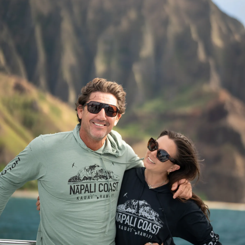 Two people in sunglasses on a boat, smiling with rocky cliffs in the background.