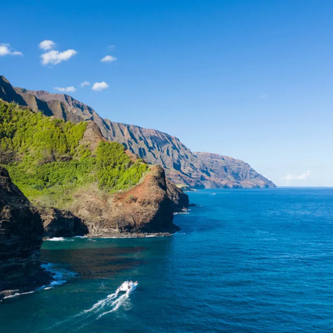 Steep cliffs with green vegetation meet blue ocean under a clear sky, a boat sails near the shore.