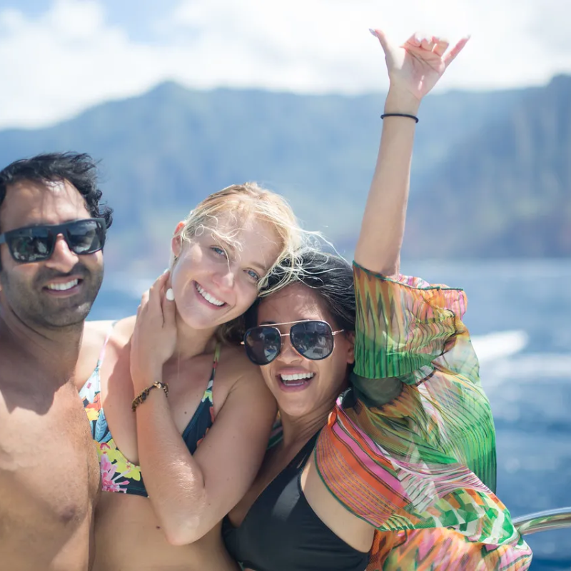 Three people in swimwear on a boat, smiling with mountains and ocean in the background.