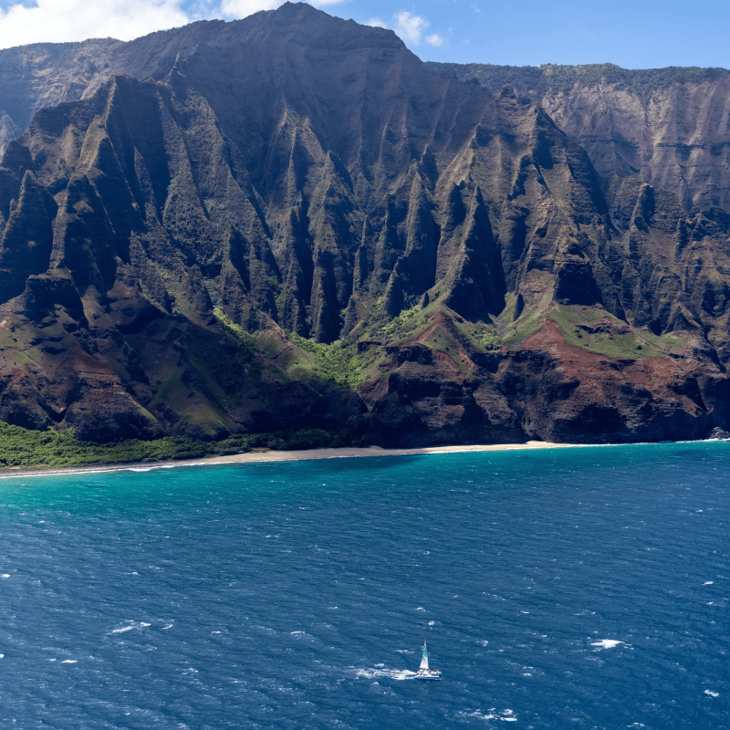 A dramatic coastline with sheer cliffs and a small sailboat on blue ocean water under a partly cloudy sky.