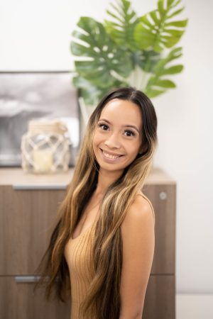 Woman with long hair smiling, standing indoors with plants in background.