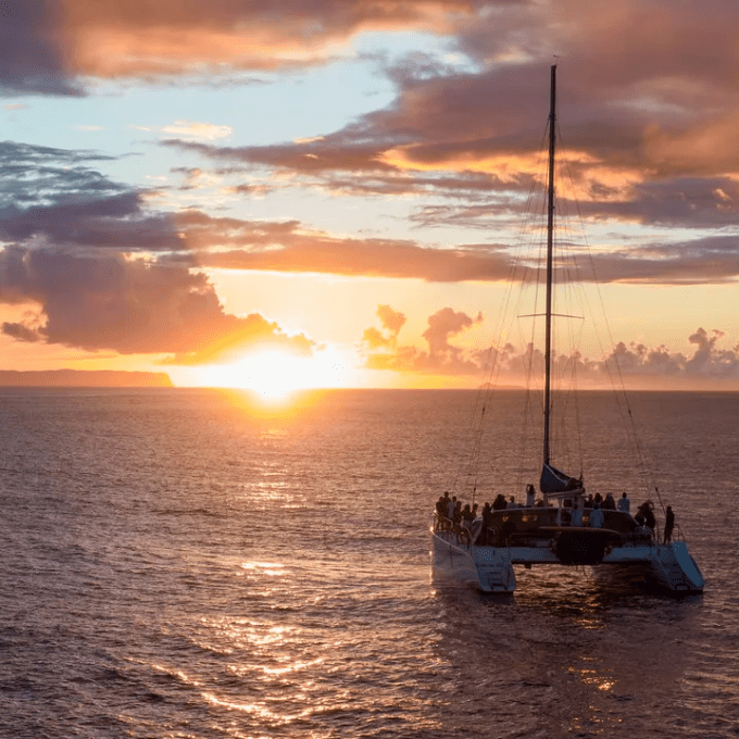 A catamaran on ocean during sunset, with vibrant clouds and horizon visible.